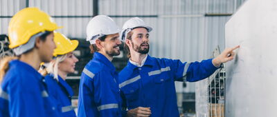 Vier Menschen in blauen Jacken und mit Helm auf dem Kopf schauen auf ein Whiteboard.